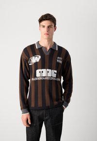 Giovane uomo che indossa un maglione a righe verticali marroni e nere con colletto grigio e la scritta "EST. 1986" e "Mads Norgaard Copenhagen" sul davanti.