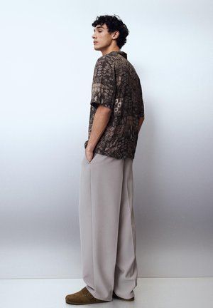 Młody mężczyzna w brązowej, wzorzystej koszuli z krótkim rękawem i szerokich beżowych spodniach stoi bokiem, z rękami w kieszeniach na jednolitym tle.