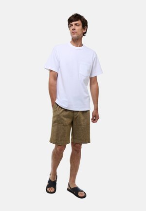 T-shirt blanc à manches courtes avec une poche sur la poitrine, associé à un short texturé beige et des sandales noires, se tenant devant un fond uni.