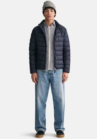 Marinblå quiltad puffjacka med dragkedja, bärs över en grårandig skjorta med knappar och ljusblå vida jeans, i kombination med svarta skor.