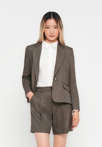 Žena so rovnými ombre vlasmi má na sebe hnedý oblek s krátkymi nohavicami a bielu košeľu s gombíkmi, stojí pred bielym pozadím.