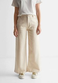 Beige široké kalhoty z bavlněného denimu, s předními kapsami, vysokým pasem a kombinované s bílými teniskami. Jednoduchý design, nezdobené.