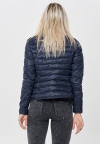 Giacca imbottita blu navy con design trapuntato, materiale in nylon testurizzato e lunghezza corta. Indossata sopra jeans grigi scuri, vista da dietro.