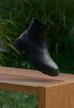 Czarne skórzane botki Chelsea z elastycznymi bocznymi panelami i gumową podeszwą, wyeksponowane w powietrzu nad drewnianym stołem, na tle zielonej trawy.