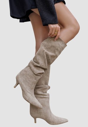 Femme portant des bottes en daim beige foncées jusqu'aux genoux avec des talons petits, ajustant une botte, vêtue d'une jupe noire sur un fond uni.