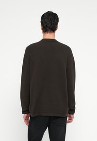 Tumši brūns teksturēts džemperis ar apaļu kakla izgriezumu un garām piedurknēm, kas izceļas ar smalku zigzaga rakstu un brīvu piegriezumu, valkāts ar tumšām džinsa biksēm.