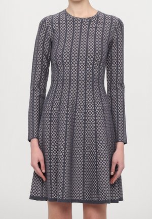 Frau trägt ein knieformendes schwarzes und graues Kleid mit langen Ärmeln und vertikalen geometrischen Mustern.