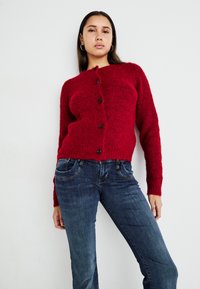 Cárdigan de punto rojo con un acabado texturizado y cinco botones negros. Combinado con unos jeans de mezclilla azul oscuro que presentan costuras visibles y bolsillos.