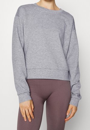 Person iført en lysegrå langærmet sweatshirt og højttaljede ribstrikkede mauve leggings, stående mod en enkel hvid baggrund.