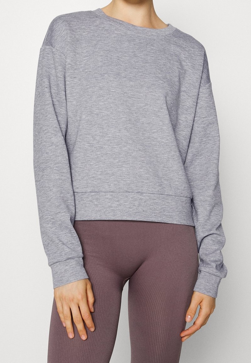 Personne portant un sweat-shirt à manches longues gris clair et un legging mauve côtelé taille haute, debout devant un fond blanc uni.