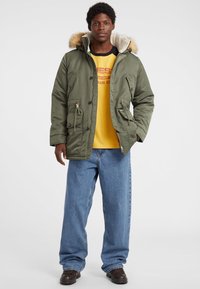 Olivgrüne Parka mit fellgefütterter Kapuze, mehreren Fronttaschen und Reißverschluss, getragen über einem gelben Hemd und hellblauen Jeans.