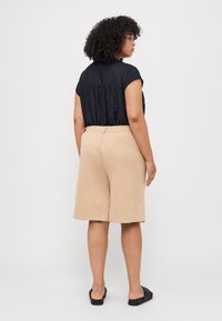 Blusa negra de manga corta con la parte trasera fruncida y pantalones cortos beige de pierna ancha, vista desde la parte trasera, combinados con sandalias negras. Fondo liso.