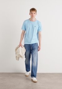 Ανοιχτό μπλε μπλούζα με στρογγυλή λαιμόκοψη και αντίθετο φιόγκο, συνδυασμένη με ξεθωριασμένο μπλε τζιν και λευκά αθλητικά παπούτσια. Το μοντέλο κρατάει ένα κρεμ μπλουζάκι.