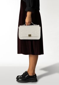 Witte gewatteerde handtas met een gouden vierkant accent, vastgehouden door een hand. Heeft een gestructureerd ontwerp en een bovenhandvat. Gedragen bij een geplooide bordeauxrode rok.