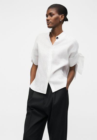 Femme à la peau foncée et aux cheveux attachés en un chignon, portant un chemisier blanc à manches courtes avec un nœud sur le côté et un pantalon noir ample, les mains dans les poches.