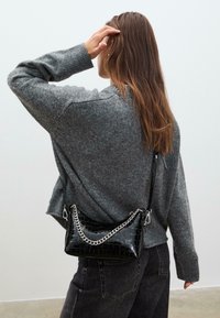 Sort håndtaske med krokodillemønster og en sølvfarvet kæderekke, bæres på tværs af kroppen over en grå uldtrøje og mørke jeans.