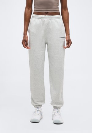 Henkilö, joka pukeutunut vaaleanharmaisiin Björn Borg -collegehousuihin, joissa on joustavat reunukset, ja valkoisiin tennareihin, seisoo valkoista taustaa vasten.