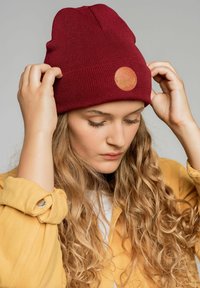 Bordowa dziana czapka typu beanie z prążkowaną teksturą i okrągłą, jasnobrązową skórzaną naszywką; noszona przez osobę z długimi, falowanymi włosami, na neutralnym tle.