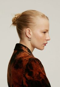 Chaqueta de pana marrón con un patrón abstracto negro, combinada con pendientes de aro plateados y de forma de corazón, mostrando un perfil lateral elegante.