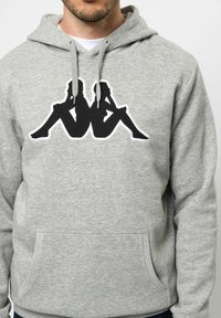 Grijze hoodie met een grote zwarte omtrek van twee zittende figuren aan de voorkant, voorzien van een kangoeroezak en een capuchon met trekkoord.