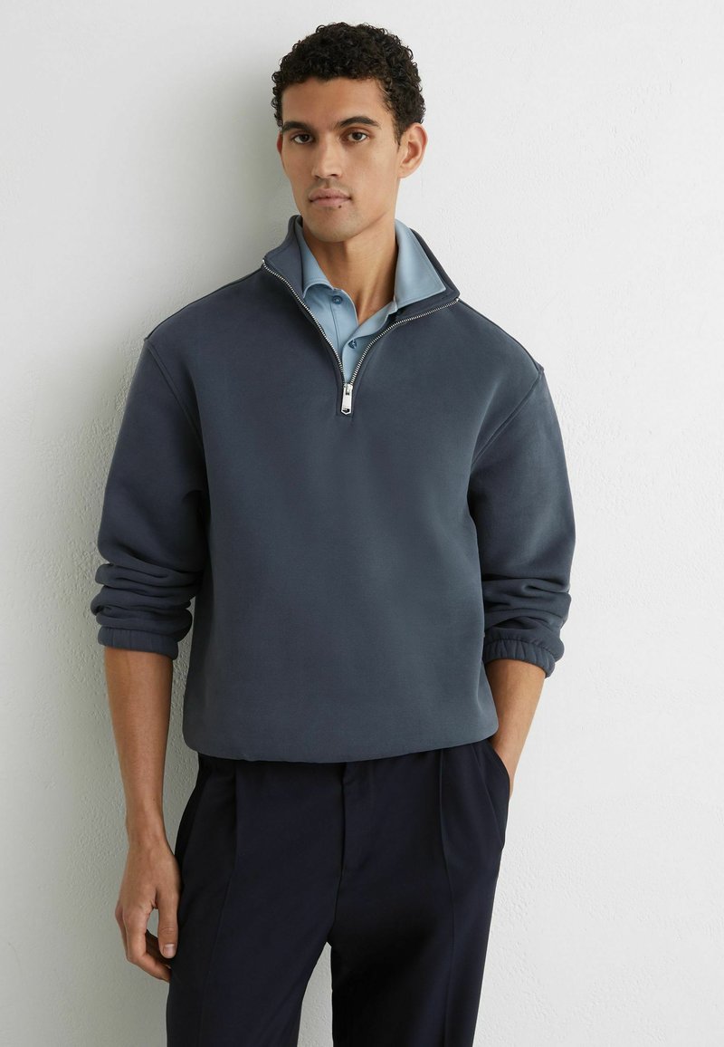 Dunkelgrauer Fleecepullover mit Viertel-Reißverschluss und gerippten Bündchen, über einem hellblauen Hemd mit Kragen getragen, mit glatter Textur und lässiger Passform.