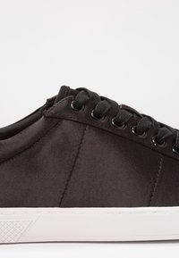 Les baskets en satin noir présentent une texture lisse, des lacets ronds et une semelle en caoutchouc blanche avec un motif en losange pour l'adhérence. La vue de côté montre des détails de couture.