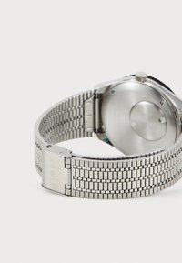 Montre en métal argenté avec un bracelet texturé, dotée d'un fermoir gravé de "TIMEX." Boîtier rond et dos lisse sans marquage visible.