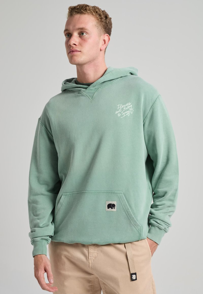 Jeune homme portant un sweat à capuche vert clair avec un texte blanc et un pantalon beige sur un fond gris uni.