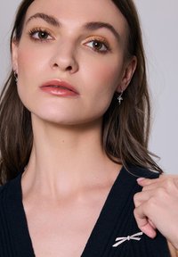 Srebrne kolczyki w kształcie kółek z małym wzorem kokardki, noszone przez modelkę z długimi brązowymi włosami, na minimalistycznym tle.