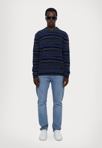 Maglione a righe blu realizzato in tessuto spesso, abbinato a jeans azzurri e sneakers bianche. Il modello indossa occhiali da sole scuri.