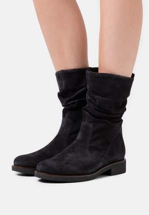 Botas de tobillo de ante negro con un diseño suelto, suela plana y ribete marrón en contraste. Textura suave y silueta simple.