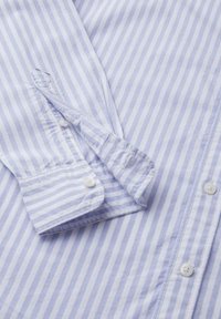Camisa de botones a rayas en azul claro y blanco, con mangas largas y puños con botones, y una textura suave. Los botones son blancos.