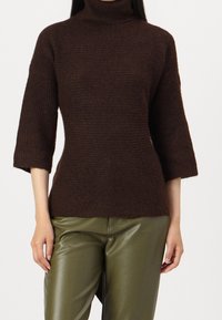 Pull-over marron ajusté avec un col roulé haut et des manches aux trois-quarts. Tissu en maille texturée, associé à un pantalon vert brillant.