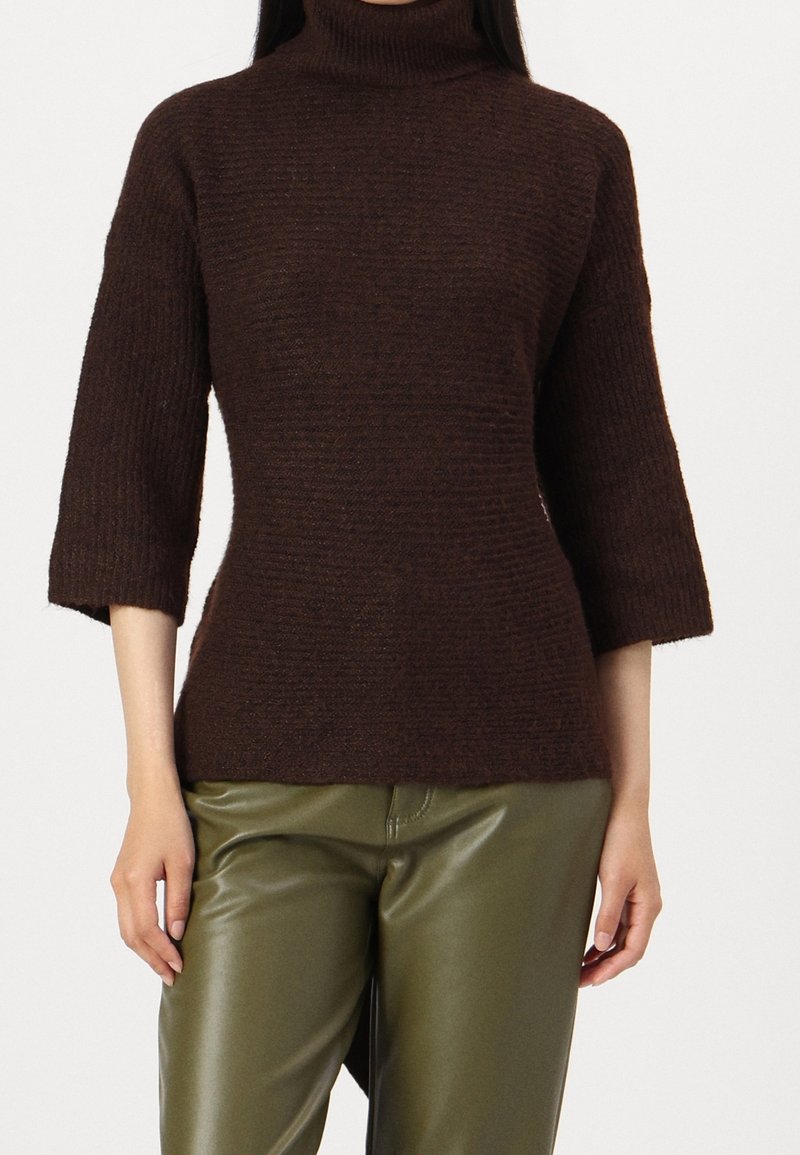 Pull-over marron ajusté avec un col roulé haut et des manches aux trois-quarts. Tissu en maille texturée, associé à un pantalon vert brillant.