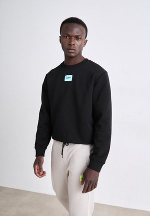 Sweatshirt noir avec l'étiquette HUGO en bleu clair, col rond et manches longues, accompagné d'un pantalon gris clair avec un accent vert.