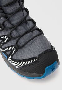 Chaussure de randonnée en textile gris avec des accents en caoutchouc noir, design rayé et semelle extérieure bleue. Comprend un système de laçage serré et un capuchon de protection pour les orteils.