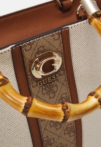 Gros plan d’un sac à main Guess beige et marron avec un logo doré « G » et des anses style bambou.