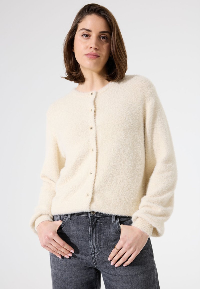 Cremefarbener, flauschiger Cardigan mit rundem Ausschnitt, Knopfleiste und langen Ärmeln. Getragen über grauen Jeans, die die weiche Textur zeigen.