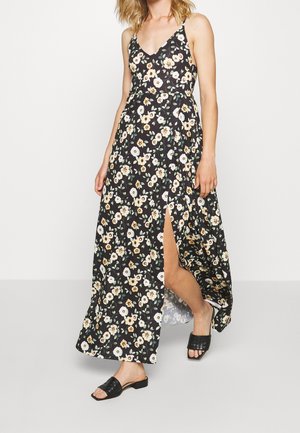 Kvinde iført en sort blomster maxi-kjole med frontslids og sorte vævede åbne tå-sandaler, stående mod en hvid baggrund.