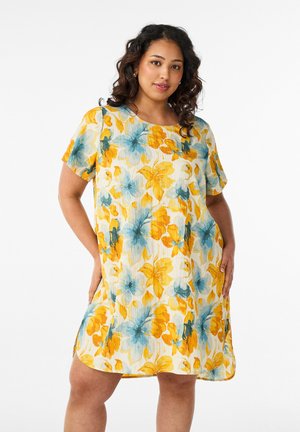 Vrouw draagt een jurk met korte mouwen tot op de knie met een geel-blauw bloemmotief, staat met de handen op de heupen.