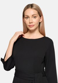 Czarna sukienka z długimi rękawami, okrągłym dekoltem i wiązaniem w talii. Wykonana z gładkiej tkaniny, z minimalnymi zdobieniami i dopasowanym krojem.