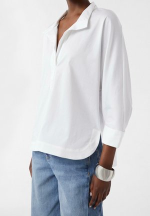 Personne portant un chemisier blanc ample à manches longues avec un col, un jean bleu et un large bracelet argenté au poignet droit.
