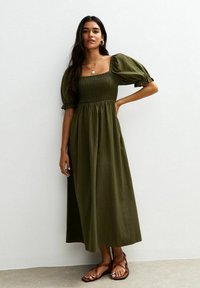 Olijfgroene maxi-jurk met een vierkante halslijn, gerimpeld lijfje en pofmouwen. Gemaakt van een lichte stof, gecombineerd met sandalen met bandjes.