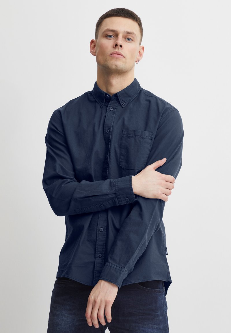 Mann med kort hår iført en mørk blå skjorte med knapper og jeans, som står med armene krysset mot en ensfarget lys bakgrunn.