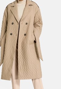 Gabardina acolchada beige con un diseño de doble botonadura, grandes solapas de muesca y botones negros. La tela tiene una textura suave.