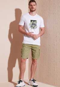 White graphic tee with green and black design, paired with light green cargo shorts. Styled with white and black sneakers. Simple background.