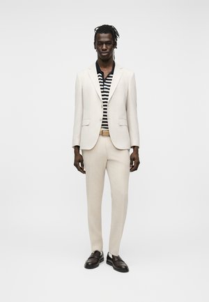 Man draagt een crème kostuum met een beige riem, een zwart-wit gestreept shirt en donkerbruine loafers, staand tegen een witte achtergrond.