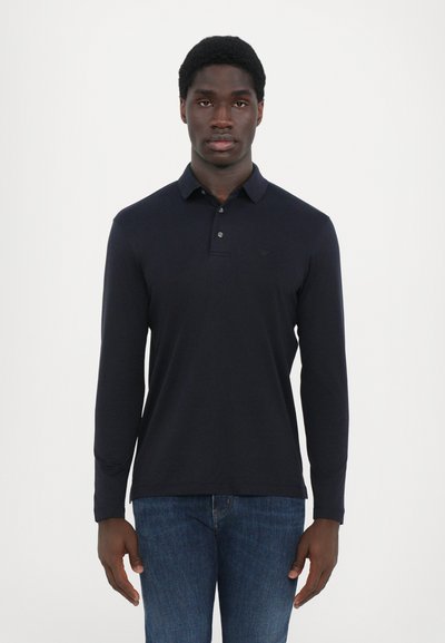 Camisa polo de manga larga en tejido liso color marino, con cuello abotonado y un sutil bordado del logo en el pecho, combinada con jeans de mezclilla.