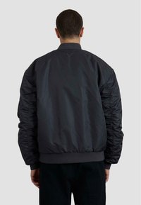 Giacca bomber nera con una texture liscia e lucida, polsini e orlo elasticizzati, con dettagli arricciati sulle maniche, vista dal retro.