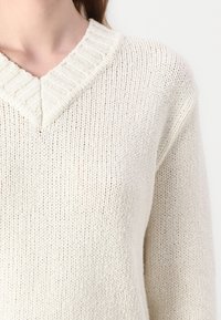 Pulover tricotat de culoare crem, cu decolteu în V, având un guler elastic și un material texturat, ușor.